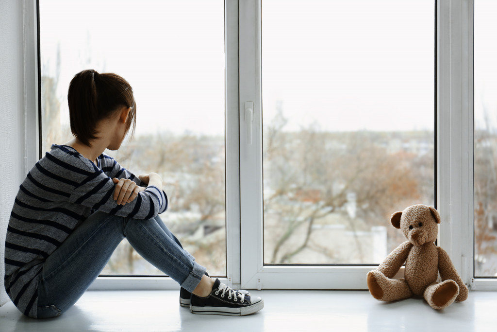 Young girl looking out a window with a bear leaning on the window.